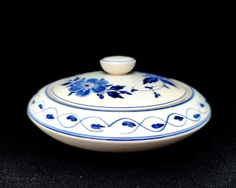 May include: A white ceramic lid with blue floral and vine patterns. The lid features a small, round knob and a blue and white wavy border. The design is reminiscent of traditional porcelain.