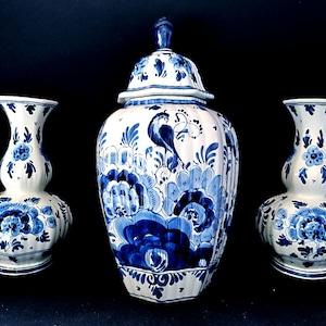 May include: A set of three ceramic vases with a blue and white floral design. The set includes a lidded jar and two smaller vases. The design features flowers, leaves, and a bird. The vases are on a black background.