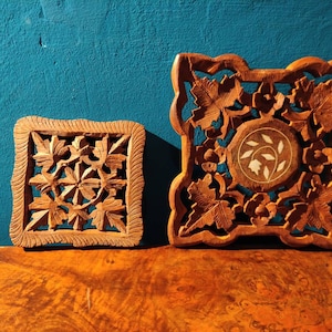 Puede incluir: Dos posavasos de madera tallados con intrincados diseños florales. El posavasos más grande presenta una incrustación blanca en el centro. Ambos posavasos son marrones y tienen un aspecto rústico.