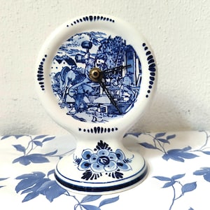 Puede incluir: Un reloj de cerámica blanca con una escena pastoril azul y blanca. El reloj tiene una esfera redonda con manecillas negras y un centro color latón. La base está decorada con motivos florales azules. El reloj se exhibe sobre una superficie con estampado floral azul y blanco.