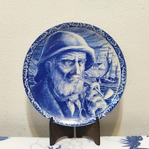 Puede incluir: Plato decorativo azul y blanco con un retrato de un hombre mayor con barba, que lleva un sombrero y fuma en pipa. El plato tiene un tema náutico, con veleros y una escena costera en el fondo.