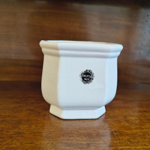 Op de afbeelding: Witte keramische plantenbak met een zeshoekige basis en een afgeronde bovenkant. De plantenbak heeft een klein, rond logo met de tekst "DELFTS WIT". De plantenbak staat op een houten oppervlak.