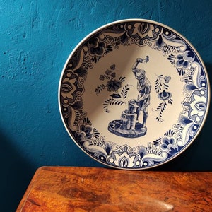 Può includere: Un piatto in ceramica bianca con un motivo floreale blu e bianco. Il centro del piatto presenta un'illustrazione blu e bianca di una persona che lavora con un martello.