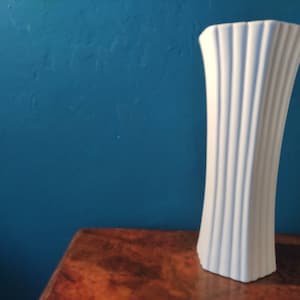 May include: White ceramic vase with vertical ridges. The vase is tall and slender, with a rounded base and a flared top.