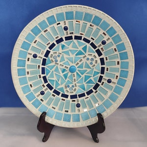 Op de afbeelding: Een blauwe en witte mozaïekplaat met een geometrisch ontwerp. De plaat staat op een zwarte standaard.