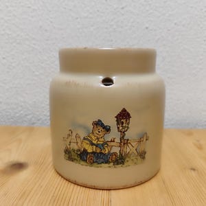 Könnte beinhalten: Eine Keramikdose mit beigefarbener Glasur und einem verspielten Design mit einem Teddybären, der eine Latzhose trägt, und einem Vogelhaus. Die Dose hat ein kleines Loch oben.