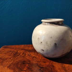 Op de afbeelding: Een witte keramische vaas met een rond lichaam en een bruine rand. De vaas heeft een gespikkelde glazuur en staat op een houten oppervlak.