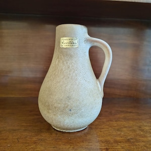 May include: A beige ceramic pitcher with a handle, featuring a textured surface and a label that reads "TONNIESHOP Carstens W-GERMANY". The pitcher is displayed on a wooden surface, showcasing its vintage design.