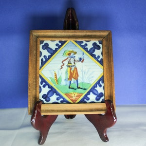May include: A framed ceramic tile with a colorful illustration of a person in a blue and yellow hat, red and yellow clothing, and holding a stick. The tile is surrounded by a blue and yellow border with a diamond pattern.