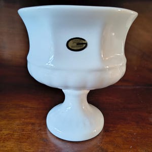 Pode incluir: Um vaso branco de cerâmica em forma de urna, com uma borda larga e canelada e uma base de pedestal. O vaso tem um acabamento liso e brilhante e um pequeno adesivo oval com texto. Uma peça decorativa.