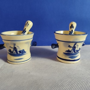 Könnte beinhalten: Zwei kleine weiße Keramik-Mörser und Stößel-Sets mit blauen und weißen Windmühlen-Designs.