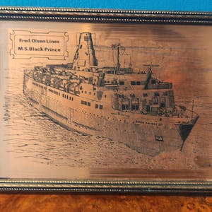 May include: A framed copper plaque with an engraved image of the M.S. Black Prince ship. The plaque is in a silver frame and has the text "Fred. Olsen Lines M.S. Black Prince" at the top.
