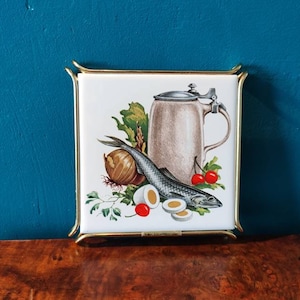Puede incluir: Una baldosa de cerámica blanca con un marco de metal dorado presenta una imagen pintada de un pez, una cebolla, huevos, cerezas y una jarra de cerveza. La baldosa es un posavasos de cocina vintage.