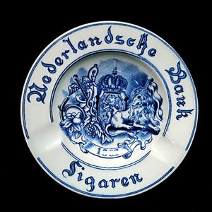 May include: A white and blue ceramic ashtray with the words "Nederlandsche Bank Sigaren" in a circular pattern. The center features a detailed blue crest with a lion, crown, and other emblems. The ashtray has a classic design.