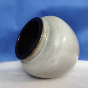 Könnte beinhalten: Eine hellgraue Keramikvase mit dunklem Rand. Die Vase hat eine abgerundete Form und eine weite Öffnung. Die Außenseite hat ein gesprenkeltes, strukturiertes Aussehen. Der Hintergrund ist blau und weiß.