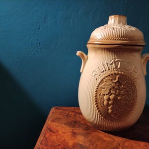 May include: A white ceramic jar with a brown lid and a grape design. The jar has the word "RUMTOPF" written on it.