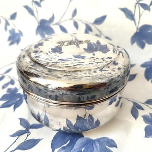 May include: A small, silver-coloured, round trinket box with a lid. The lid is inscribed with the word "Times" and features a floral design. The box is set against a white background with blue floral patterns.