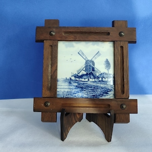 May include: A wooden framed tile featuring a blue and white windmill scene. The tile is set in a dark brown wooden frame with a built-in stand. The artwork depicts a traditional Dutch windmill, houses, and a tree.