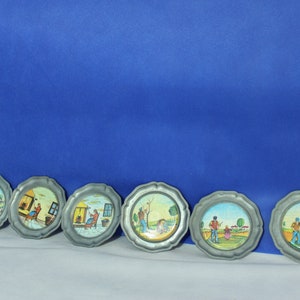 May include: Set of six round pewter coasters with painted scenes of people in a rural setting. The coasters feature a variety of scenes, including a man working in a field, a woman sitting on a porch, and a family enjoying a picnic.