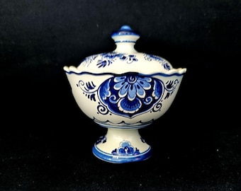 May include: A white and blue ceramic lidded bowl with a pedestal base. The bowl and lid are decorated with blue floral and scrollwork designs. The bowl has a scalloped edge and a small knob on top of the lid.