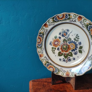 May include: A white ceramic plate with a blue and orange floral design. The plate is on a wooden stand.
