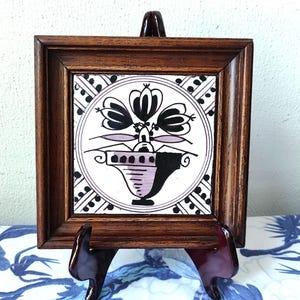 May include: Square framed tile with a floral design in a dark wood frame. The tile features a black and white floral arrangement in a vase with lavender accents. The frame is displayed on a wooden easel.