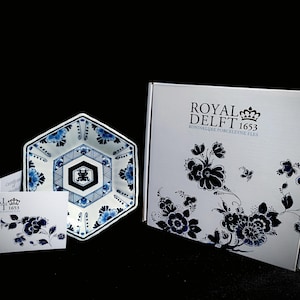 Może przedstawiać: Biała, sześciokątna miska ceramiczna z niebiesko-białym wzorem kwiatowym. Miska ma czarne i białe centrum ze stylizowanym chińskim znakiem. Miska jest zapakowana w białe pudełko z niebiesko-białym wzorem kwiatowym i tekstem "Royal Delft 1653 Koninklijke Porceleijne Flies".