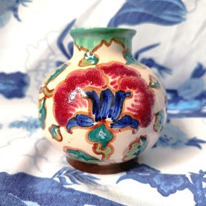 Könnte beinhalten: Eine kleine, runde Keramikvase mit weißem Hintergrund und bunten Blumenmustern. Die Vase hat einen grünen Rand und ein rotes, blaues und grünes Blumenmuster.