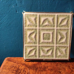 Puede incluir: Un azulejo de cerámica blanco con un patrón geométrico verde y dorado, enmarcado en un marco de metal dorado. El azulejo es una posavasos decorativo.