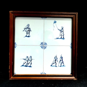 May include: A framed artwork featuring four white tiles with blue illustrations of figures engaged in various activities. The artwork is framed in a dark brown wooden frame. The illustrations depict people playing and interacting.