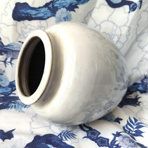 Könnte beinhalten: Eine weiße Keramikvase mit rundem Körper und einer breiten Öffnung. Die Vase hat eine dezente Glasur und eine leicht strukturierte Oberfläche.