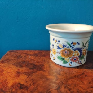 Könnte beinhalten: Weißer Keramiktopf mit floralem Design in Blautönen, Gelb, Orange und Grün. Der Topf hat eine runde Form und einen leicht ausgestellten Rand.