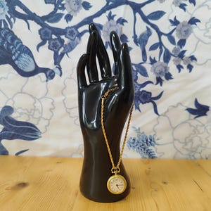 Könnte beinhalten: Ein schwarzer, handförmiger Ausstellungsständer hält eine goldfarbene Taschenuhr an einer Kette. Die Uhr hat ein weißes Zifferblatt mit schwarzen Ziffern. Der Hintergrund zeigt ein blau-weißes Blumenmuster.
