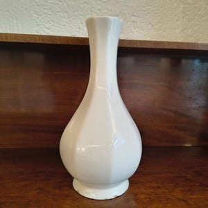 May include: A white ceramic vase with a bulbous body and a slender, faceted neck. The vase has a glossy finish and sits on a small, circular base. The background is a dark wooden surface.