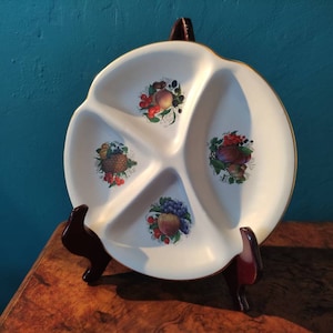 Puede incluir: Un plato de servir de cerámica blanca con cuatro secciones, cada una decorada con un diseño de fruta diferente. El plato tiene un borde dorado y está sobre un soporte de madera.