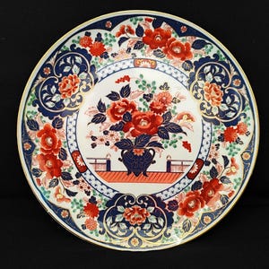 Puede incluir: Un plato de cerámica blanca con un borde azul y dorado. El plato presenta un diseño floral con flores rojas, azules y verdes. El centro del plato tiene un arreglo floral en un jarrón azul.