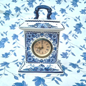 Op de afbeelding: Een witte en blauwe keramische klok met een handvat. De wijzerplaat is goudkleurig met zwarte Romeinse cijfers. De klok is versierd met blauwe bloempatronen en een blauw handvat. De klok is een decoratief item.