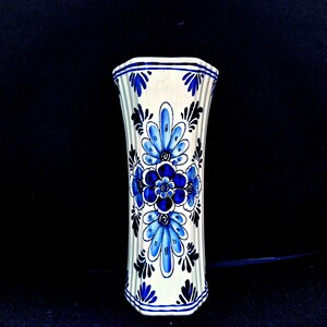 Può includere: Un vaso in ceramica bianca con un motivo floreale blu. Il vaso ha un corpo scanalato e una parte superiore e inferiore svasata. Il motivo floreale presenta fiori e foglie blu stilizzati. Il vaso è alto circa 20 cm.