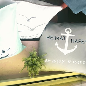 Könnte beinhalten: Mehrere dekorative Kissen mit maritimen Motiven. Ein graues Kissen zeigt einen weißen Anker und die Worte "Heimat Hafen" mit Koordinaten. Andere Kissen haben Möwen- und Kompassdesigns. Die Kissen sind in verschiedenen Blau- und Weißtönen gehalten.