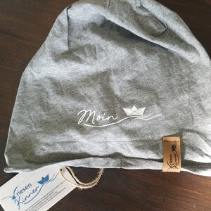 Könnte beinhalten: Graue Mütze mit einem weißen gestickten Design eines Papierboots und dem Wort "Moin". Die Mütze hat ein braunes Leder-Etikett mit dem Text "riesen kinner" in Schwarz.
