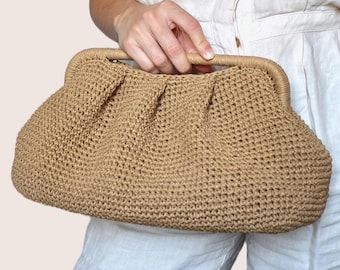 May include: A tan crochet clutch bag with a gathered top. The bag is held by a person wearing a white linen shirt and pants, and a straw hat. The bag's texture and color suggest a natural, summery style.