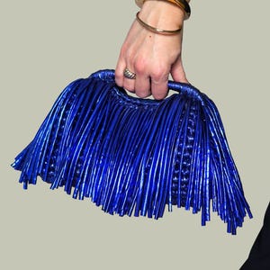 May include: A vibrant blue clutch with a fringe design. The bag is held by a person wearing gold bracelets and a ring. The clutch has a handle and a textured surface, adding a touch of elegance to the accessory.