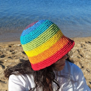 May include: A rainbow crochet bucket hat with a wide brim. The hat is made with a variety of colors, including red, orange, yellow, green, blue, and purple. The hat is sitting on a person's head, and the person is wearing a white shirt.