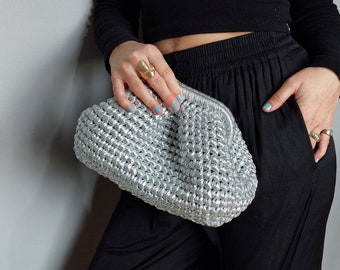Może przedstawiać: Srebrna torebka typu clutch wykonana na szydełku z marszczonym wzorem. Torebkę trzyma osoba ubrana w czarne spodnie i czarną bluzkę.
