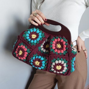 Op de afbeelding: Een gehaakte handtas met een houten handvat. De tas is gemaakt van kleurrijke vierkanten met een bloemenpatroon in tinten rood, roze, geel, blauw en groen.