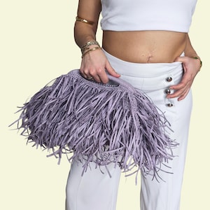 May include: A lavender fringed handbag with a rounded handle. The bag is held against a background of white trousers and a white top. The fringe is long and creates a textured effect.
