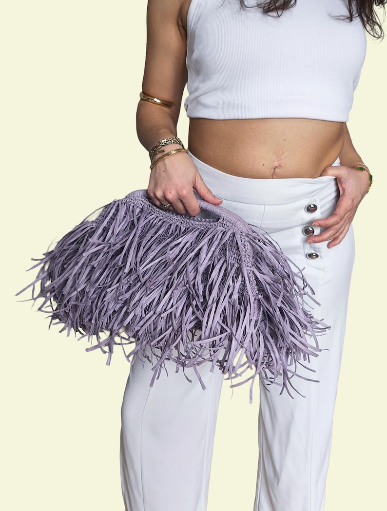 May include: A lavender fringed handbag with a rounded handle. The bag is held against a background of white trousers and a white top. The fringe is long and creates a textured effect.