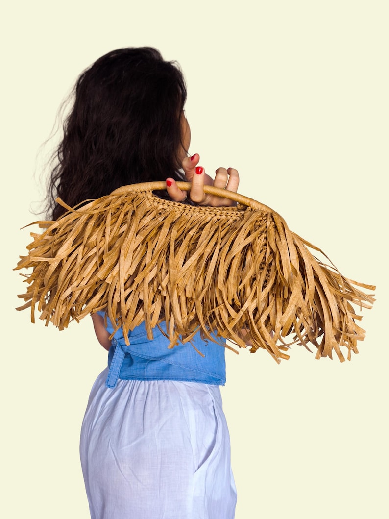 May include: A woven straw handbag with long, fringed edges. The bag is a light brown colour and has a handle. The bag is held by a person wearing a blue top and white trousers. The background is a solid, light yellow colour.