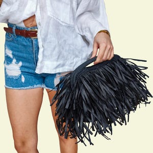 May include: A black fringed handbag with a short handle. The bag is held by a person wearing a white shirt and blue denim shorts. The bag's fringe is made of long, thin strips of material.