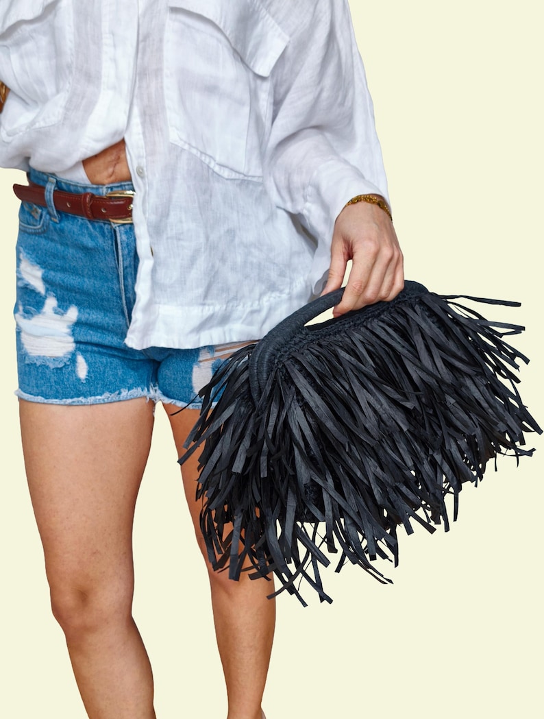 May include: A black fringed handbag with a short handle. The bag is held by a person wearing a white shirt and blue denim shorts. The bag's fringe is made of long, thin strips of material.